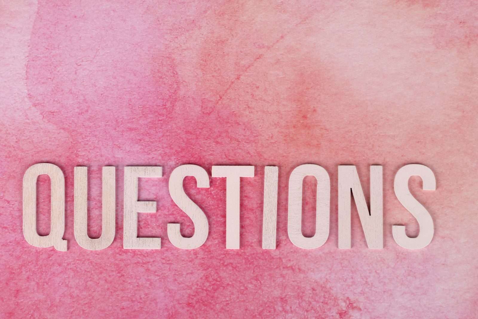 Wooden 'QUESTIONS' text on pink watercolor background, perfect for design and art concepts.