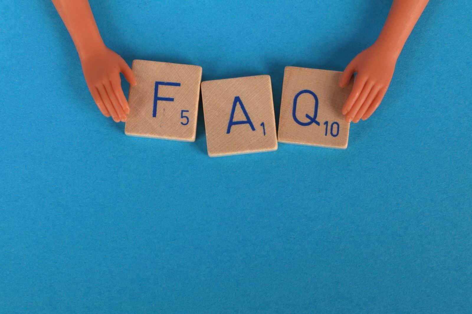 Flat lay of scrabble tiles spelling 'FAQ' with toy hands on a blue background, creating a conceptual image.